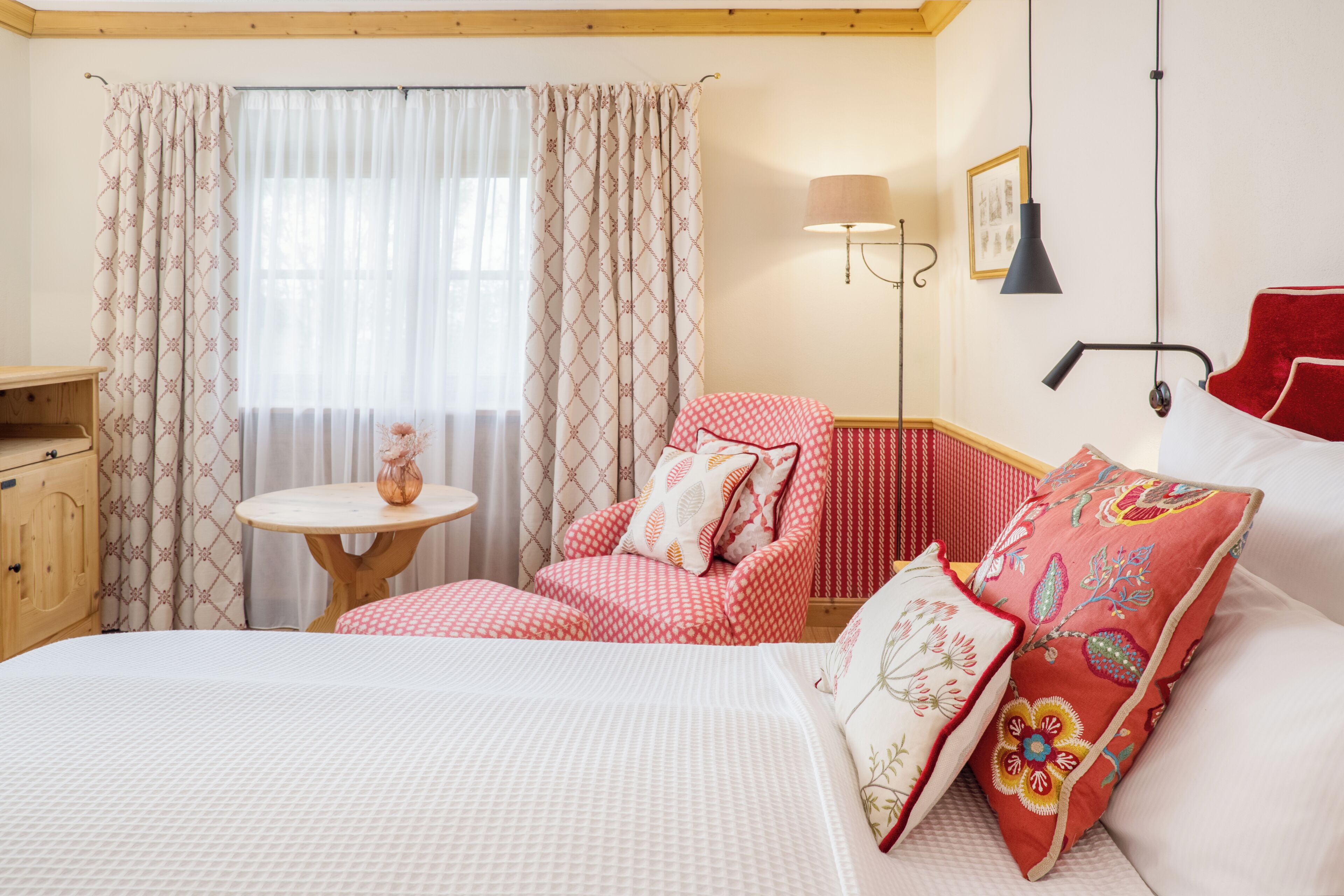 A hotel room with a bed, a red and white patterned armchair, a small round table with a vase, and a lamp on the wall.