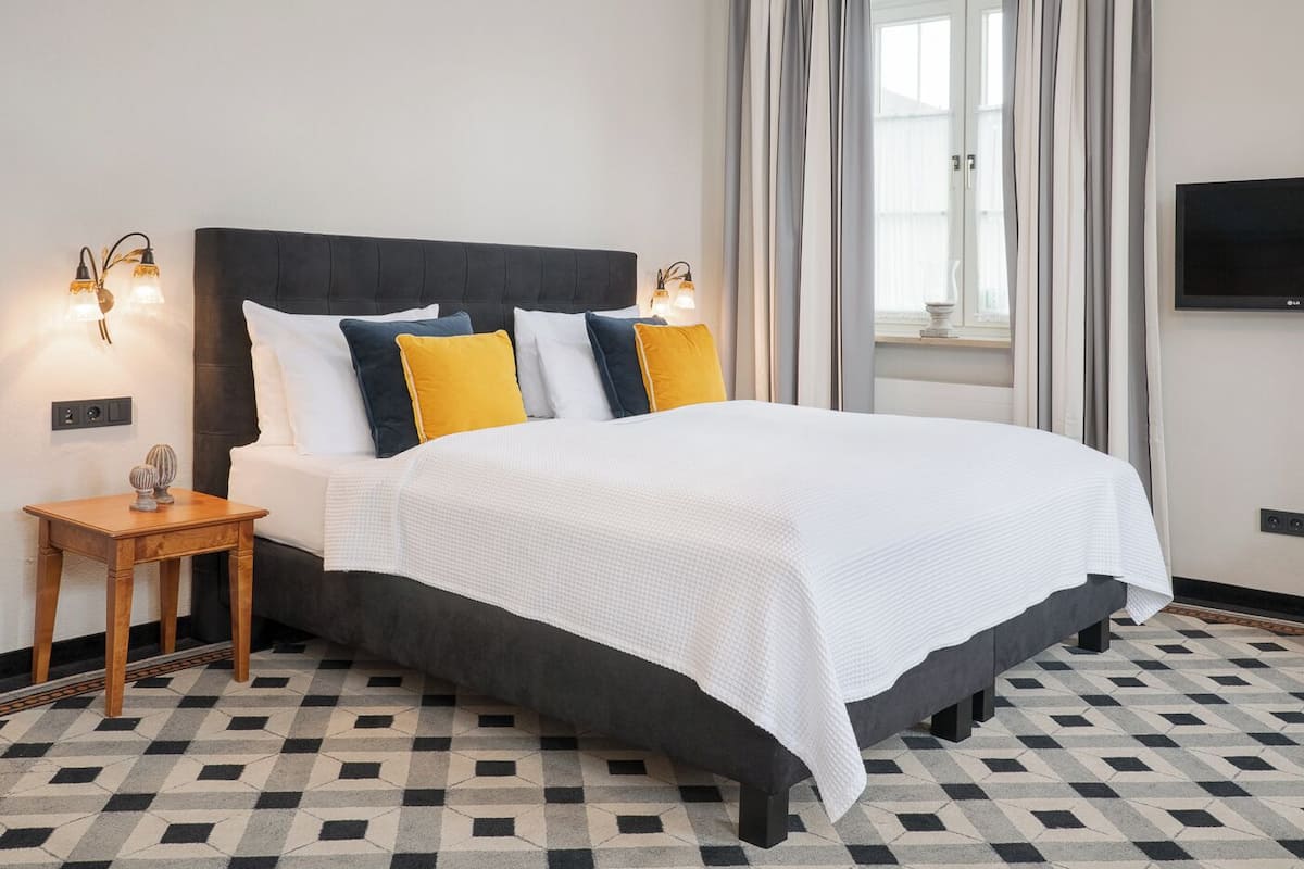Una cama con cabecera oscura, ropa de cama blanca y varios almohadones amarillos y azules. Junto a la cama, una mesita de noche de madera con objetos decorativos. El cuarto tiene piso de baldosas con un diseño geométrico y un ventanal con cortinas.