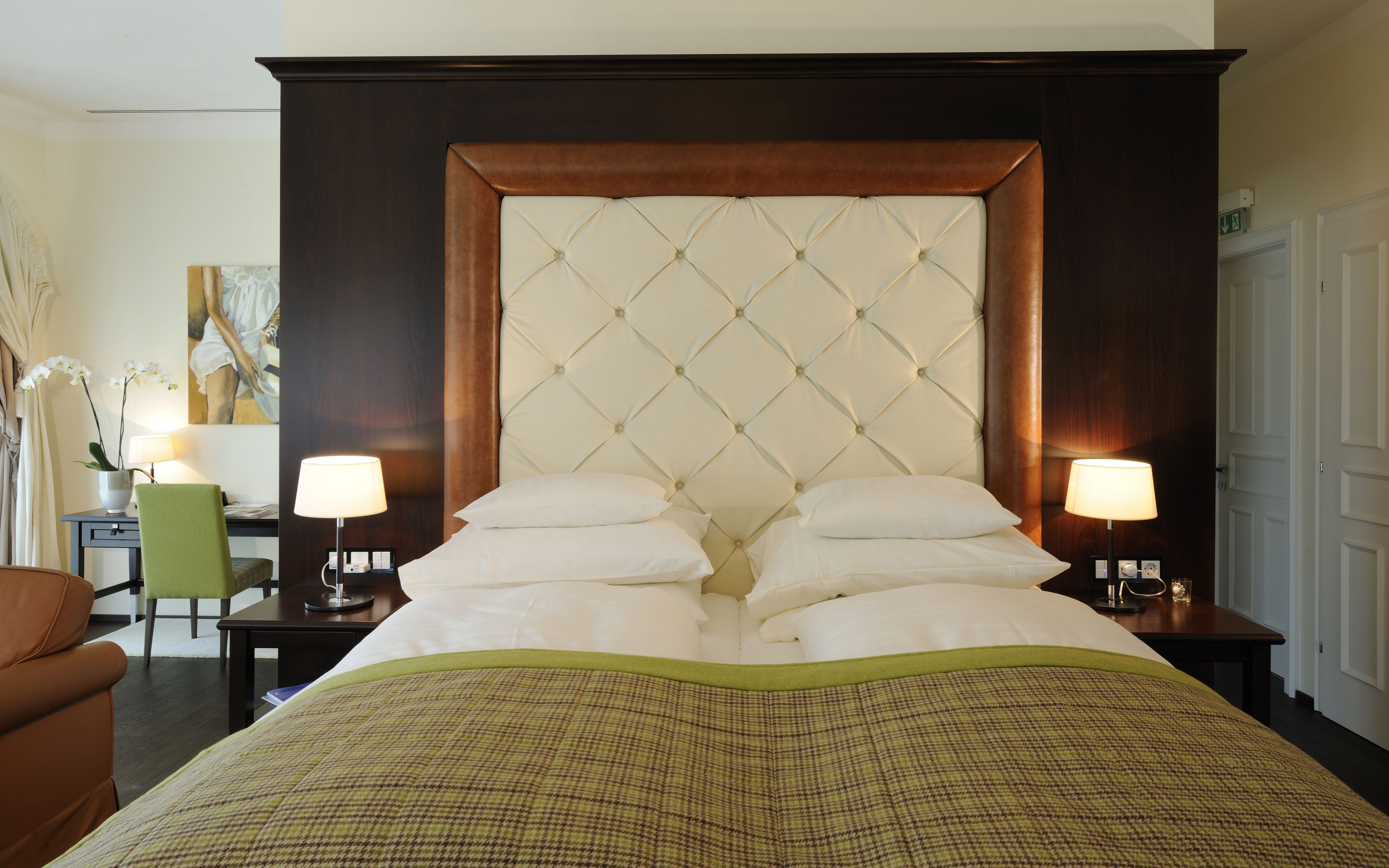Habitación de hotel con una cama grande, dos mesitas de noche con lámparas, una silla verde, un escritorio y un cuadro en la pared.