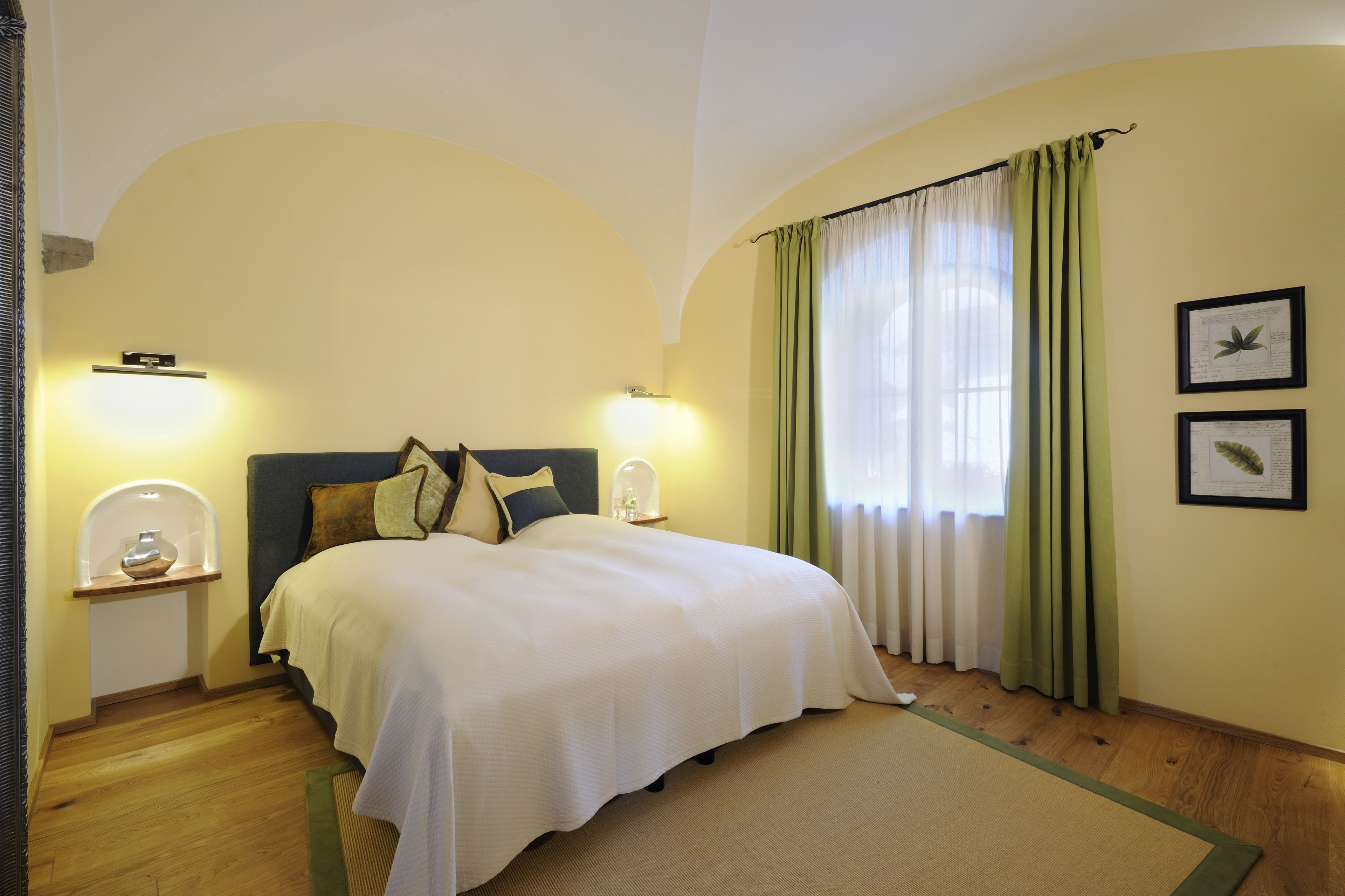 A bedroom with a large bed, two bedside tables, a nightstand, a window with green curtains, and two framed pictures on the wall.
