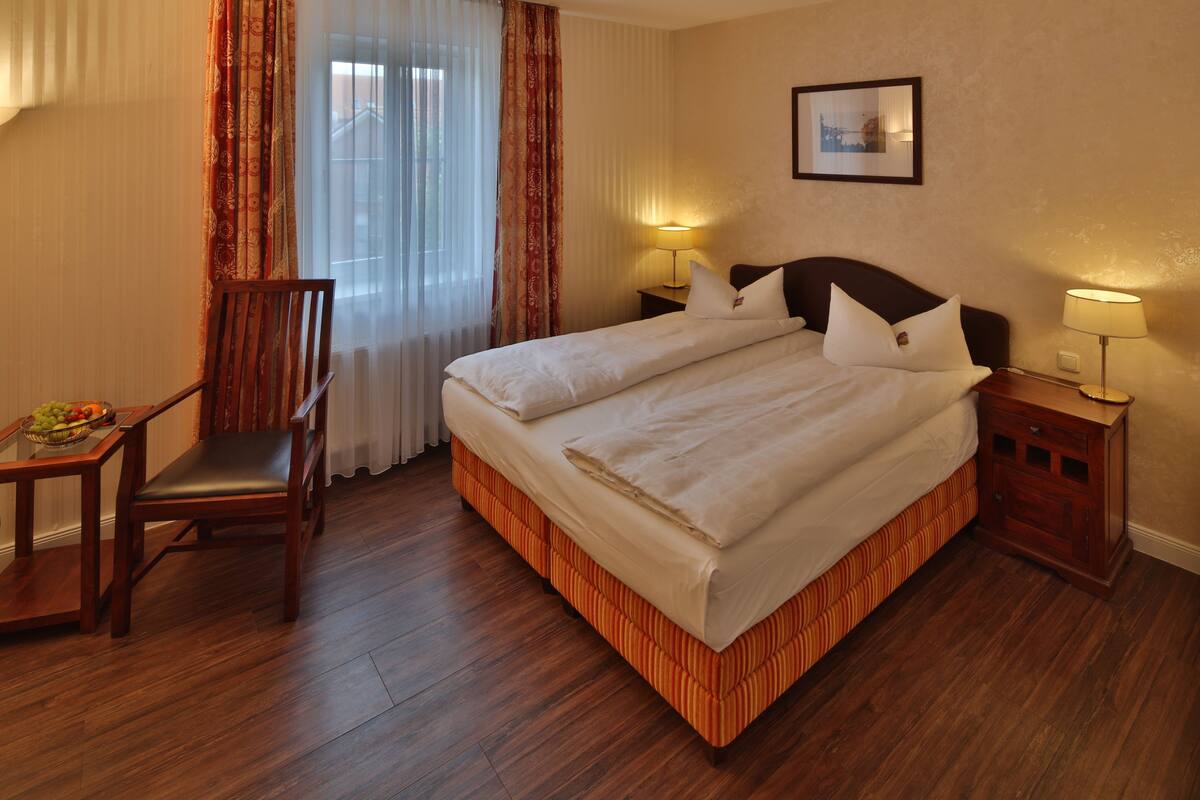 A hotel room with a double bed, two bedside tables with lamps, a wooden chair, a small table with fruit, and a window with sheer curtains.
