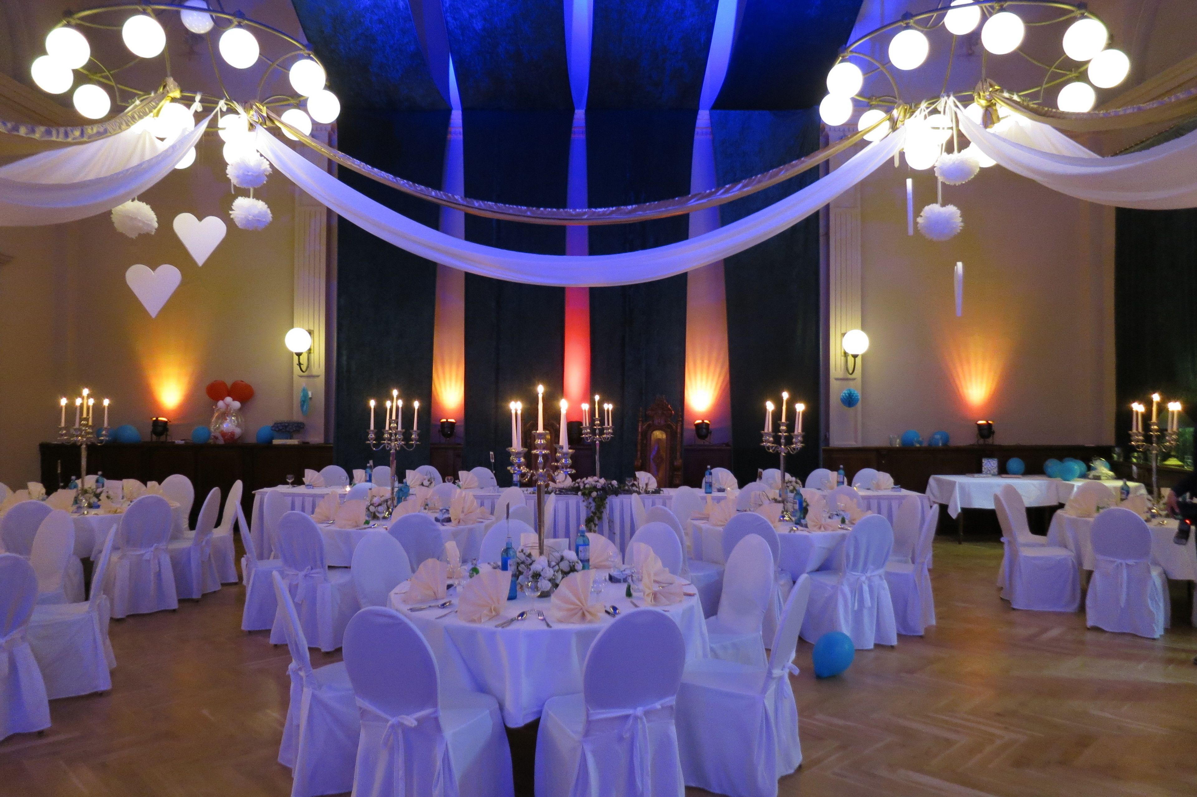 Un salón de banquetes con mesas redondas preparadas para un evento formal, cubiertas con tela blanca y globos azules.