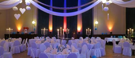Un salón de banquetes con mesas redondas preparadas para un evento formal, cubiertas con tela blanca y globos azules.