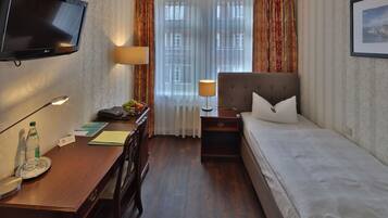 Una habitación de hotel con una cama, un escritorio con teléfono, televisión, lámpara y ventana con cortinas.