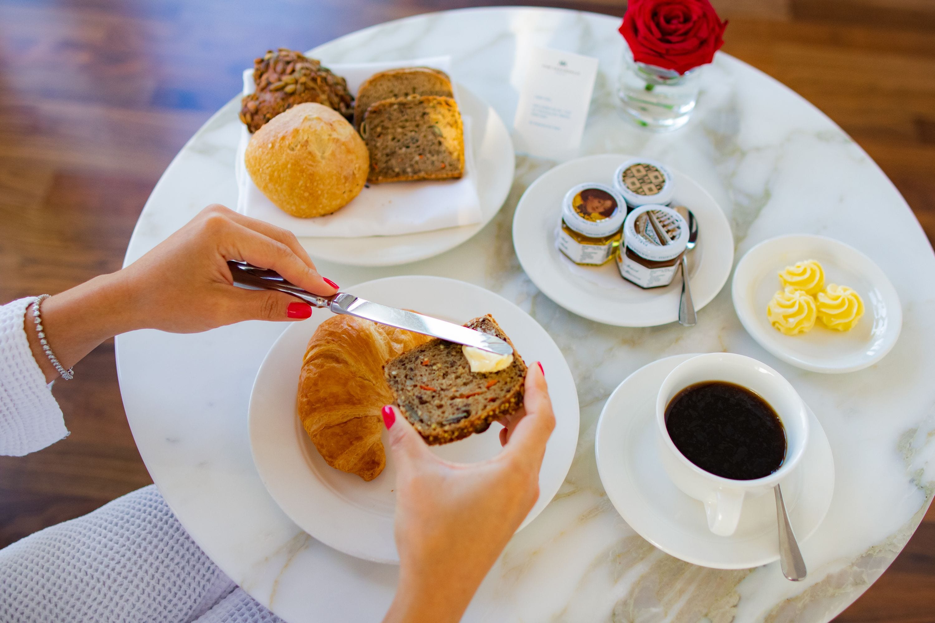 Una mesa con un croissant, una rebanada de pan, un frasco pequeño de mermelada, una taza de café y un plato con pasteles.