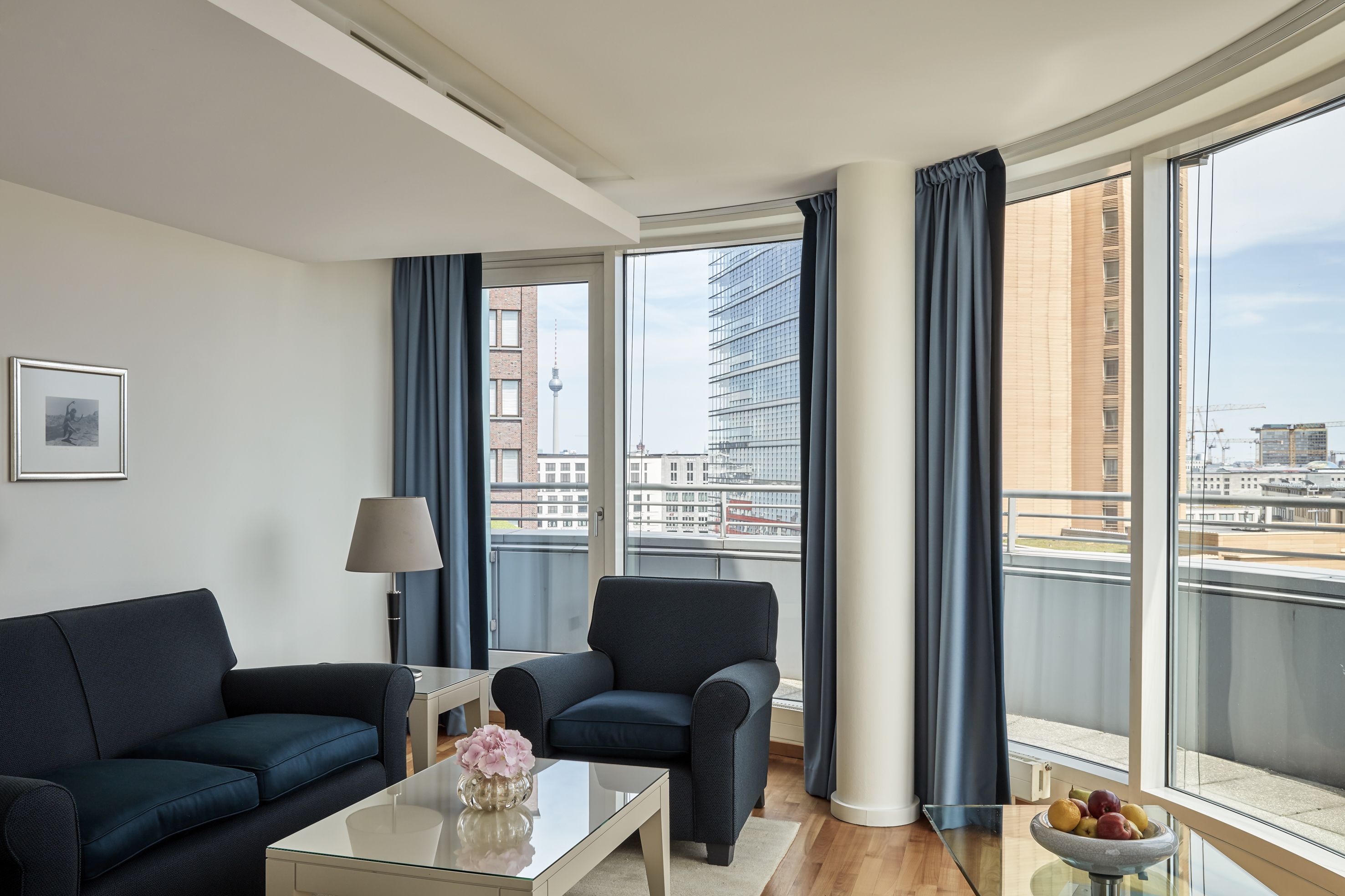 Una sala de estar moderna con sofá, sillón y mesa de centro, ventanas amplias con cortinas y vistas a edificios de la ciudad.