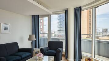Una sala de estar moderna con sofá, sillón y mesa de centro, ventanas amplias con cortinas y vistas a edificios de la ciudad.