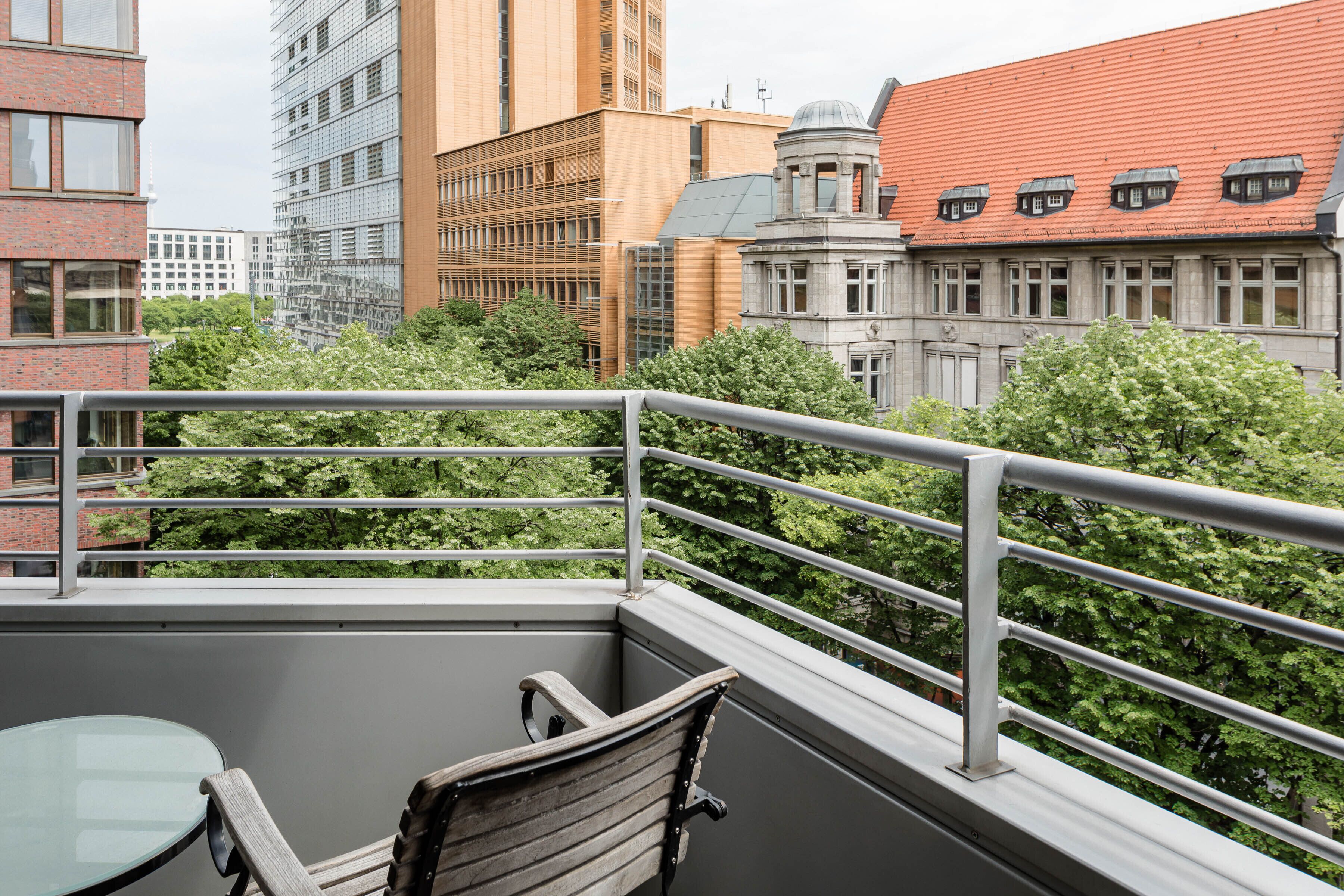 A balcony with a glass table and wooden chair, overlooking a cityscape with various buildings and trees.