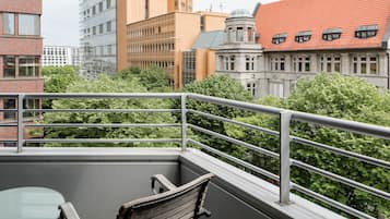 A balcony with a glass table and wooden chair, overlooking a cityscape with various buildings and trees.