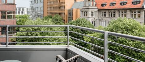 A balcony with a glass table and wooden chair, overlooking a cityscape with various buildings and trees.