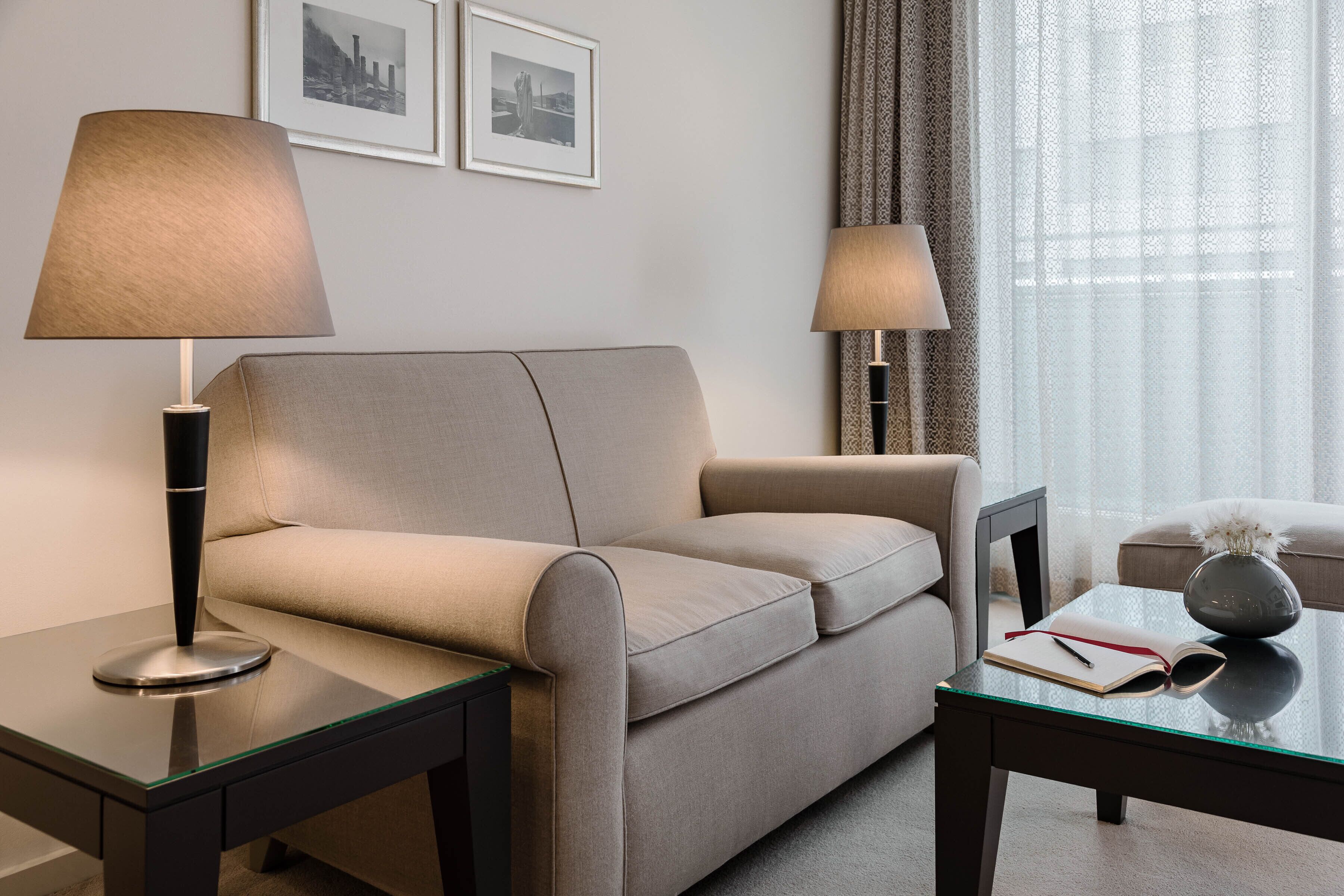 A modern hotel room with a beige sofa, two side tables with lamps, a glass coffee table, and a framed picture on the wall.