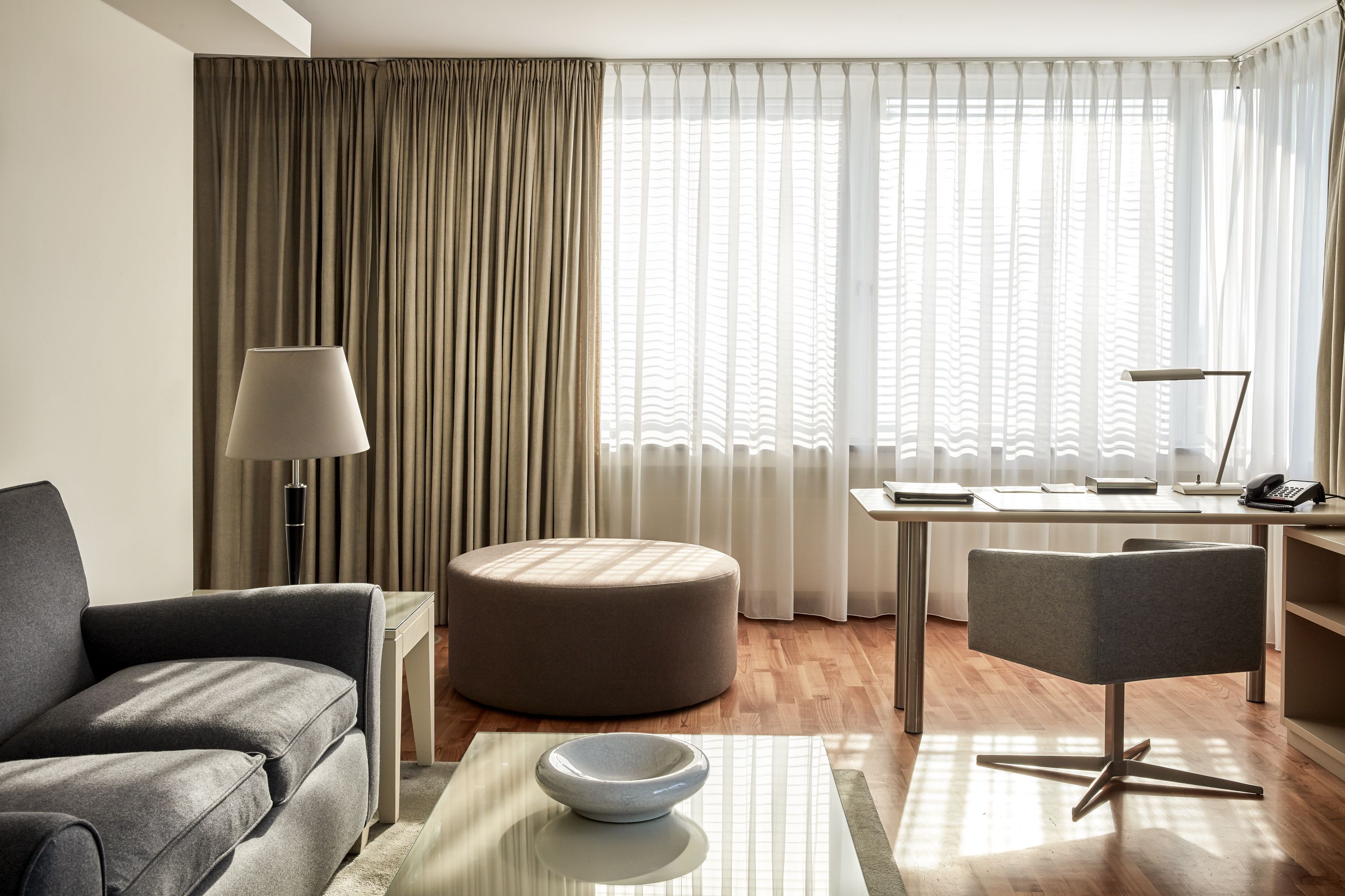 A modern hotel room with a grey sofa, a round ottoman, a desk with a lamp, and a view through sheer curtains.
