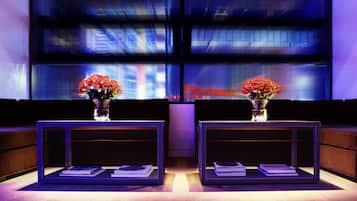 En moderne lobby med to glasborde, hvert med en vase blomster og bøger. Store vinduer giver udsigt over bybilledet.