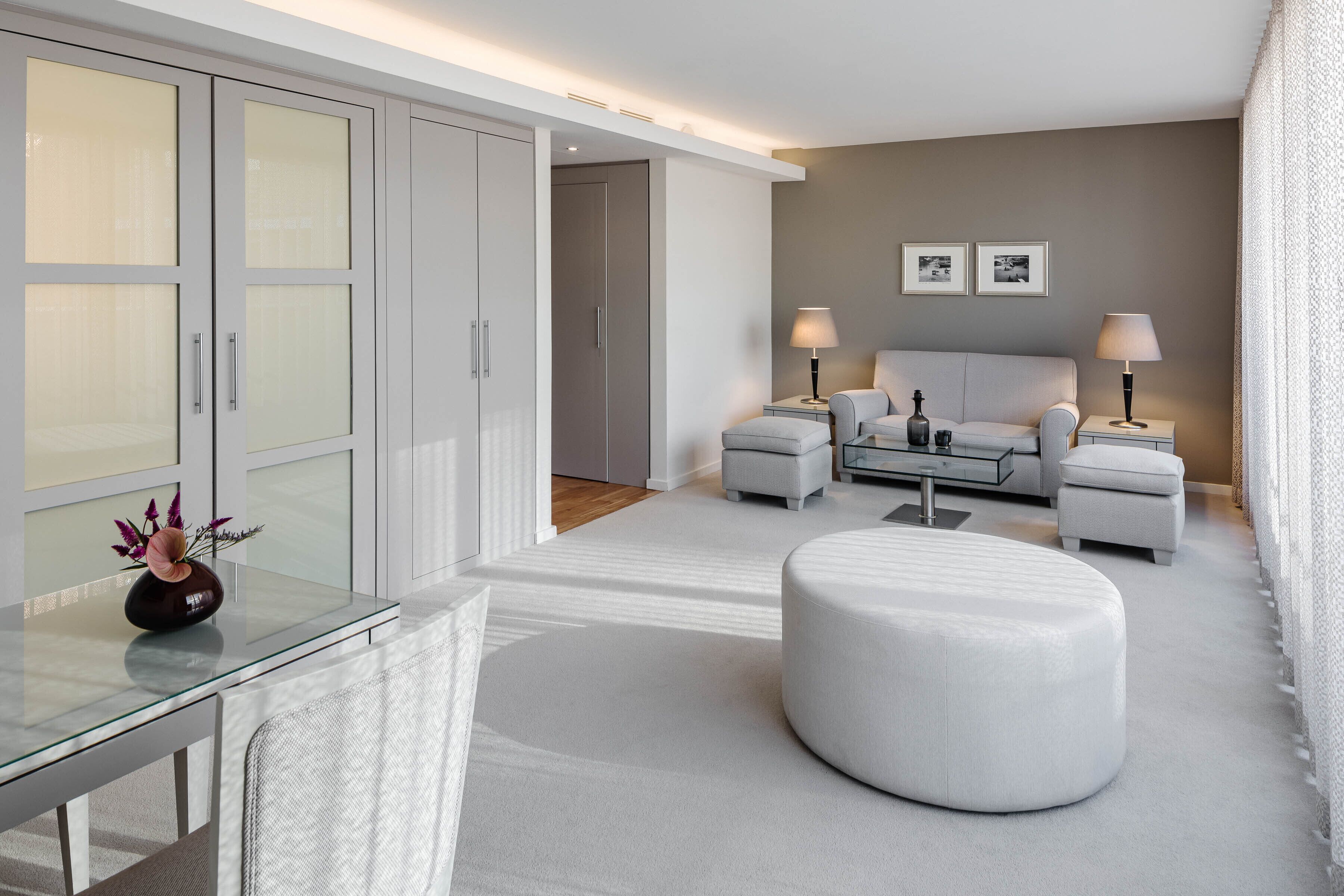 A modern hotel room with a glass dining table, a round ottoman, a sofa, two bedside tables with lamps, and a wardrobe with frosted glass doors.