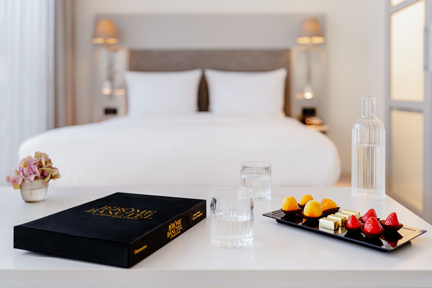 Habitación de hotel con una cama bien hecha, una mesita de noche con un libro, vasos y una bandeja con diversos postres.