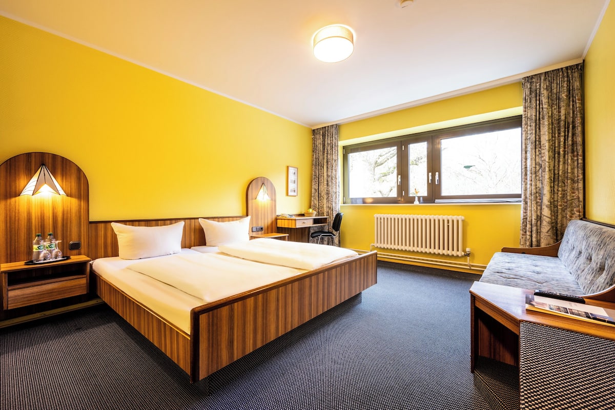 A hotel room with a wooden bed, a desk, a chair, a lamp, and a window with curtains.