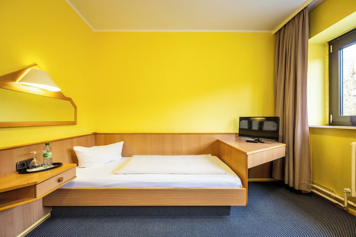 A compact hotel room with a single bed, a bedside table with a lamp, a TV, and a window with curtains.