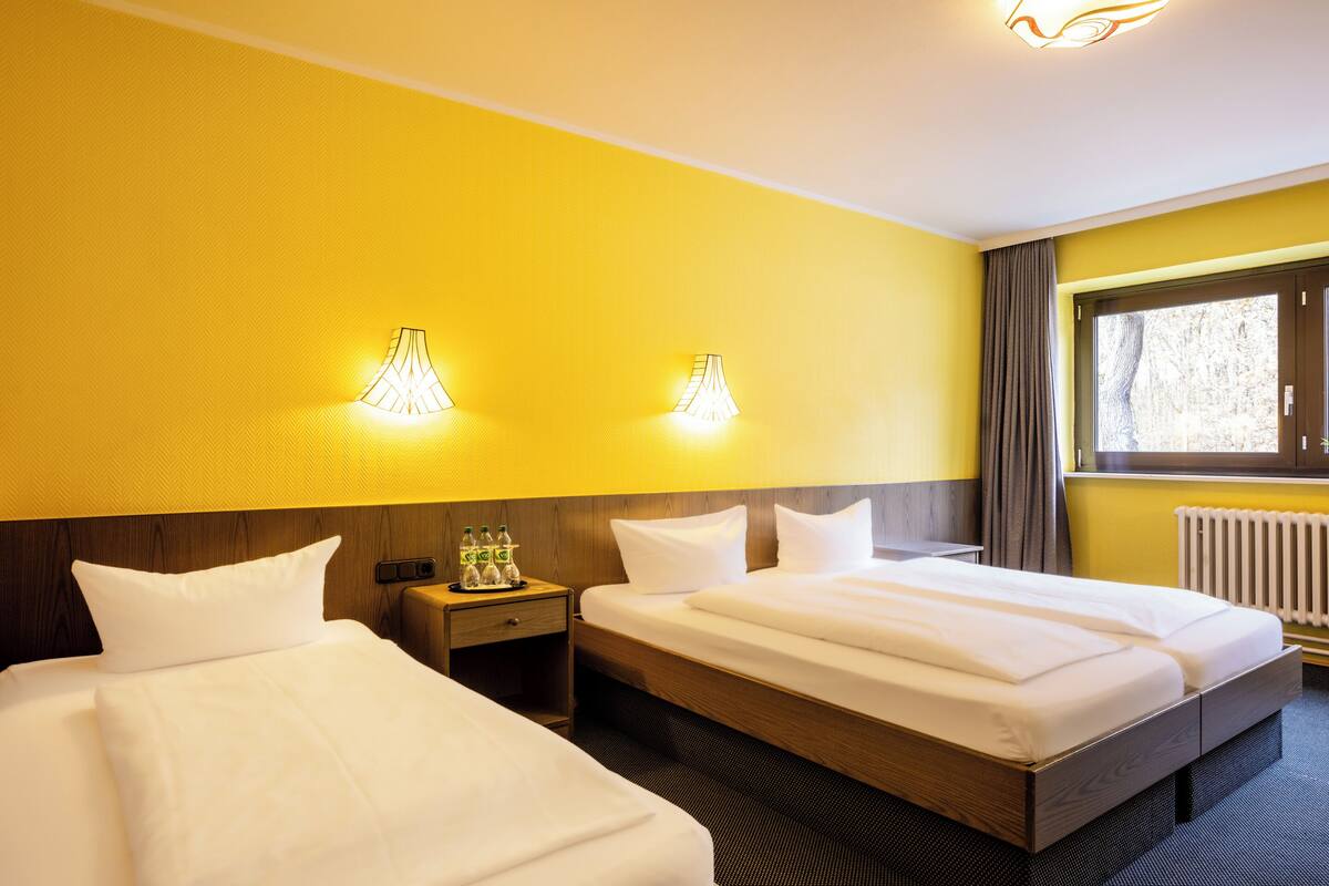 A hotel room with two beds, a wooden headboard, a nightstand with a bottle of water, and a window with curtains.
