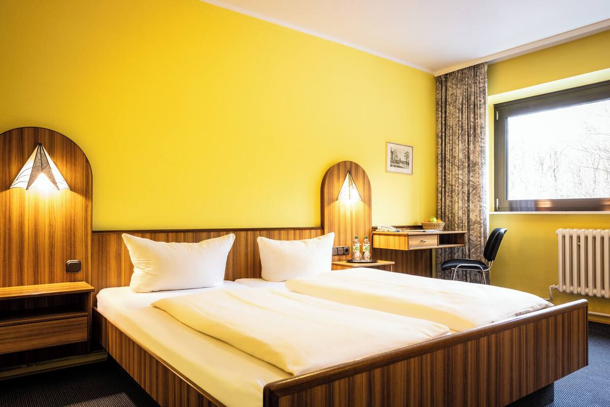 A hotel room with a wooden bed, two bedside tables, a desk with a chair, a window with curtains, and a radiator.