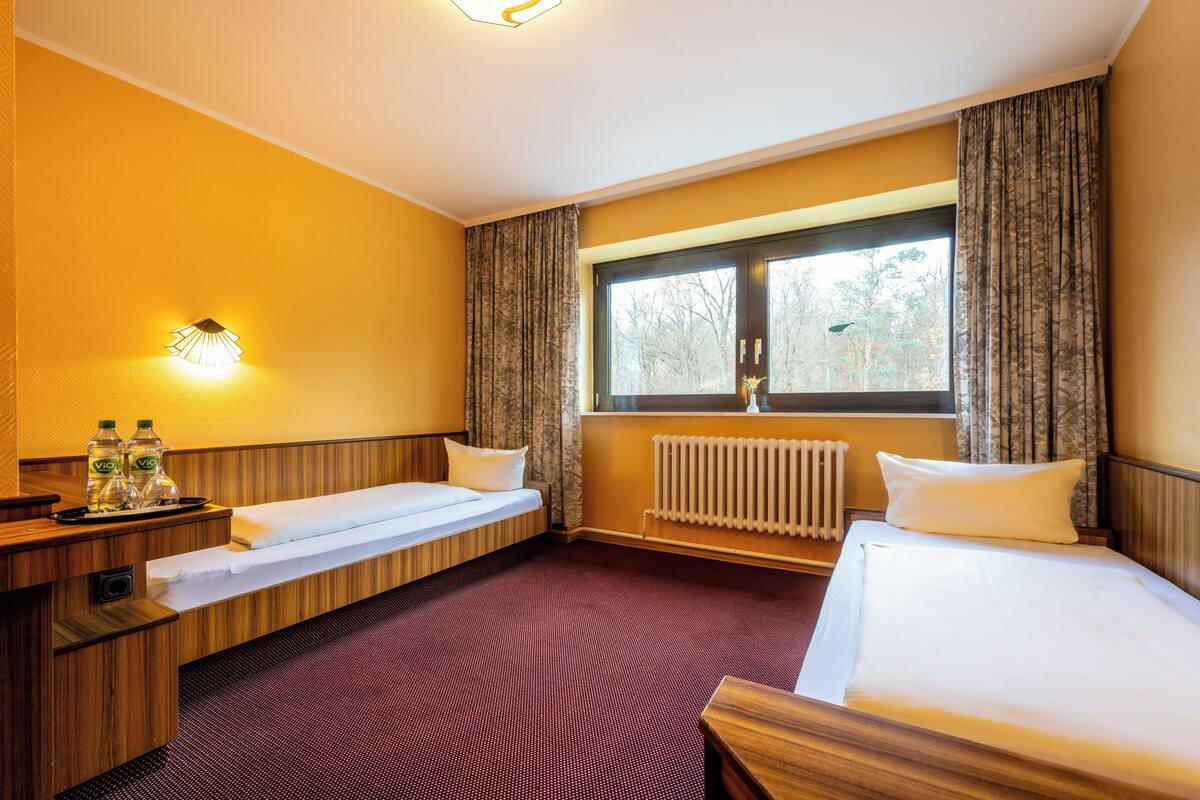 A hotel room with a bed, a bench, a bedside table with bottles, a window with curtains, and a radiator.
