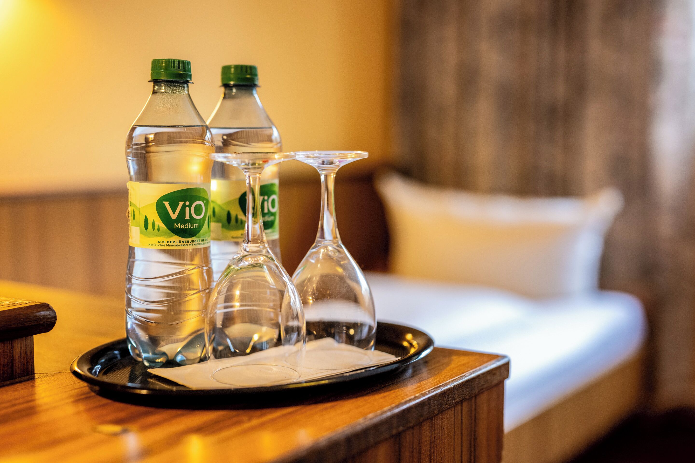 A tray with two bottles of Vio Medium water and two clear glasses on a wooden table in a hotel room.