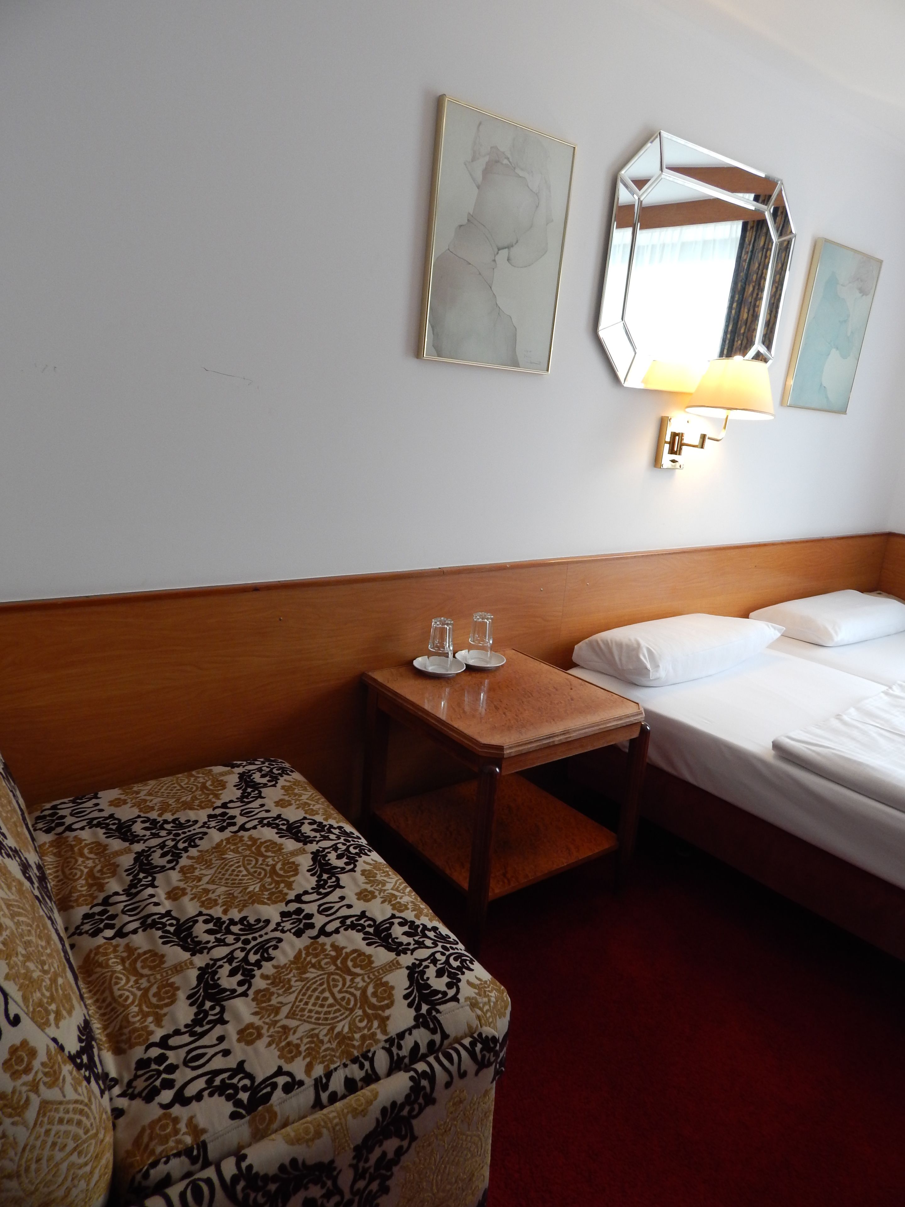 Habitación de hotel con cama, cabecera de madera, mesita de noche con dos vasos, sofá estampado y dos espejos colgados en la pared.