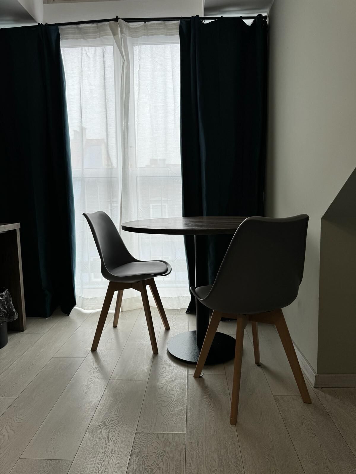 A modern room with a round table and two chairs, large windows with dark curtains, and a light-colored wooden floor.