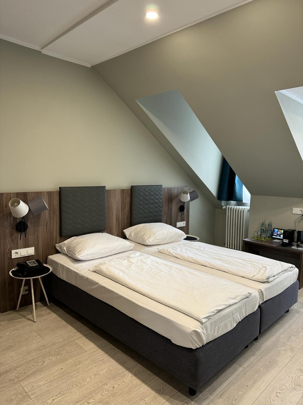 A modern bedroom with a large bed, two bedside tables, a desk with a computer, and a view of the sky through a skylight.