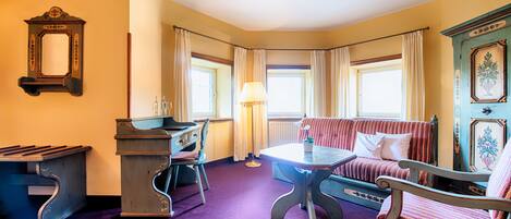 A room with a purple carpet, a desk with a chair, a table, a sofa, and a window with curtains.
