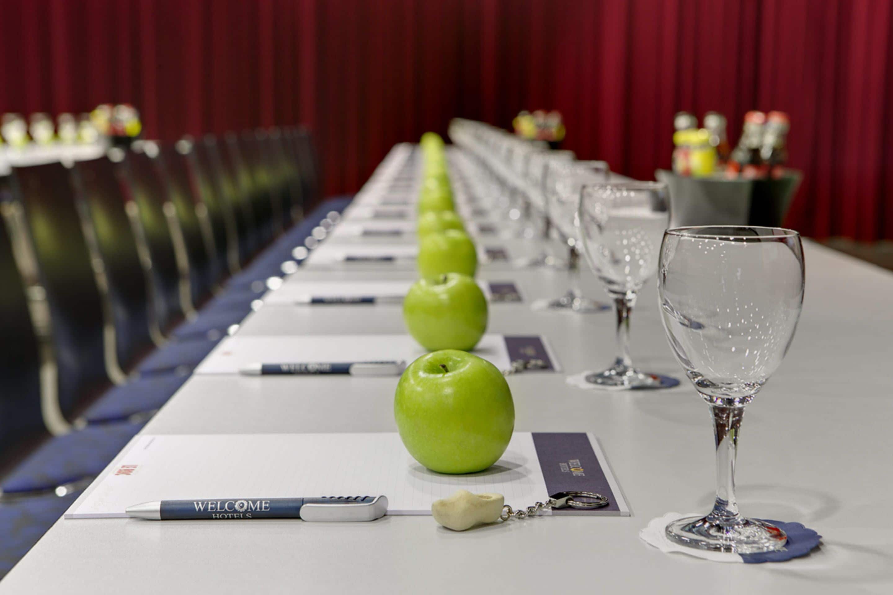 Una mesa de conferencias con una hilera de manzanas verdes, un bolígrafo, un llavero y una copa de vino.