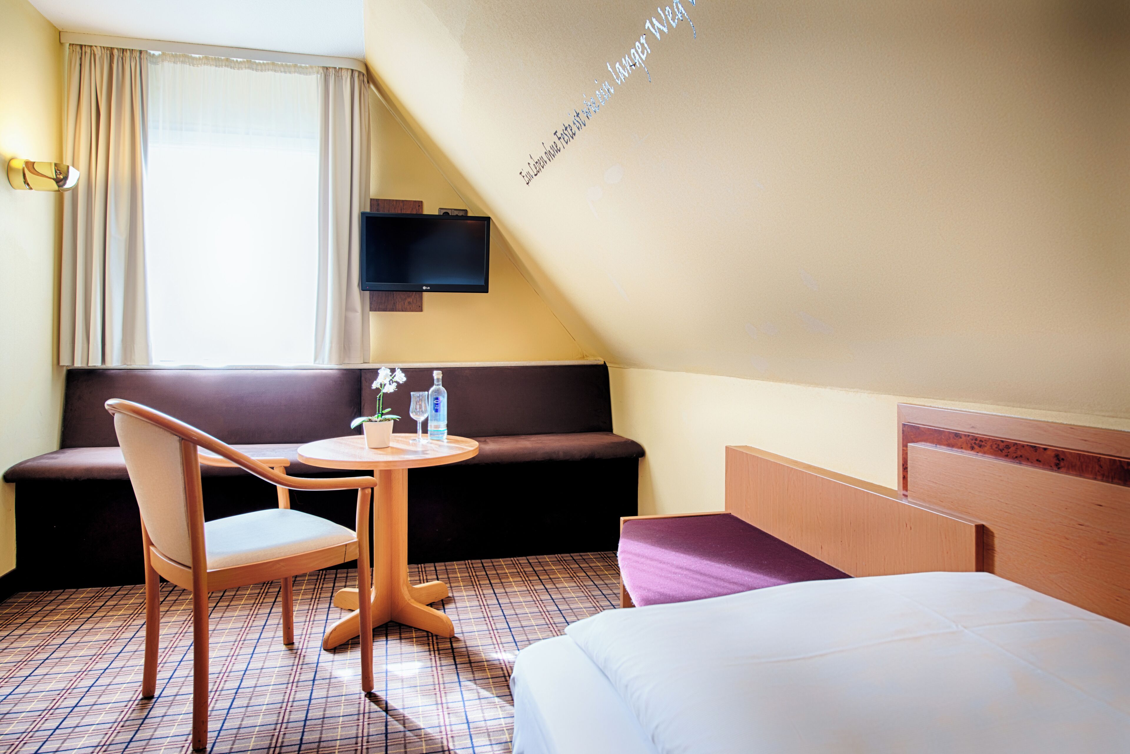 Een hotelkamer met een bed, een bank, een tafel met een plant en een fles, en een aan de muur bevestigde televisie.