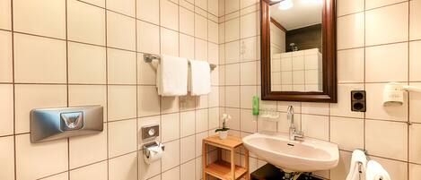 A modern bathroom with a large mirror, a sink, and a toilet. There are two towels hanging on the wall and a small wooden shelf with a plant.