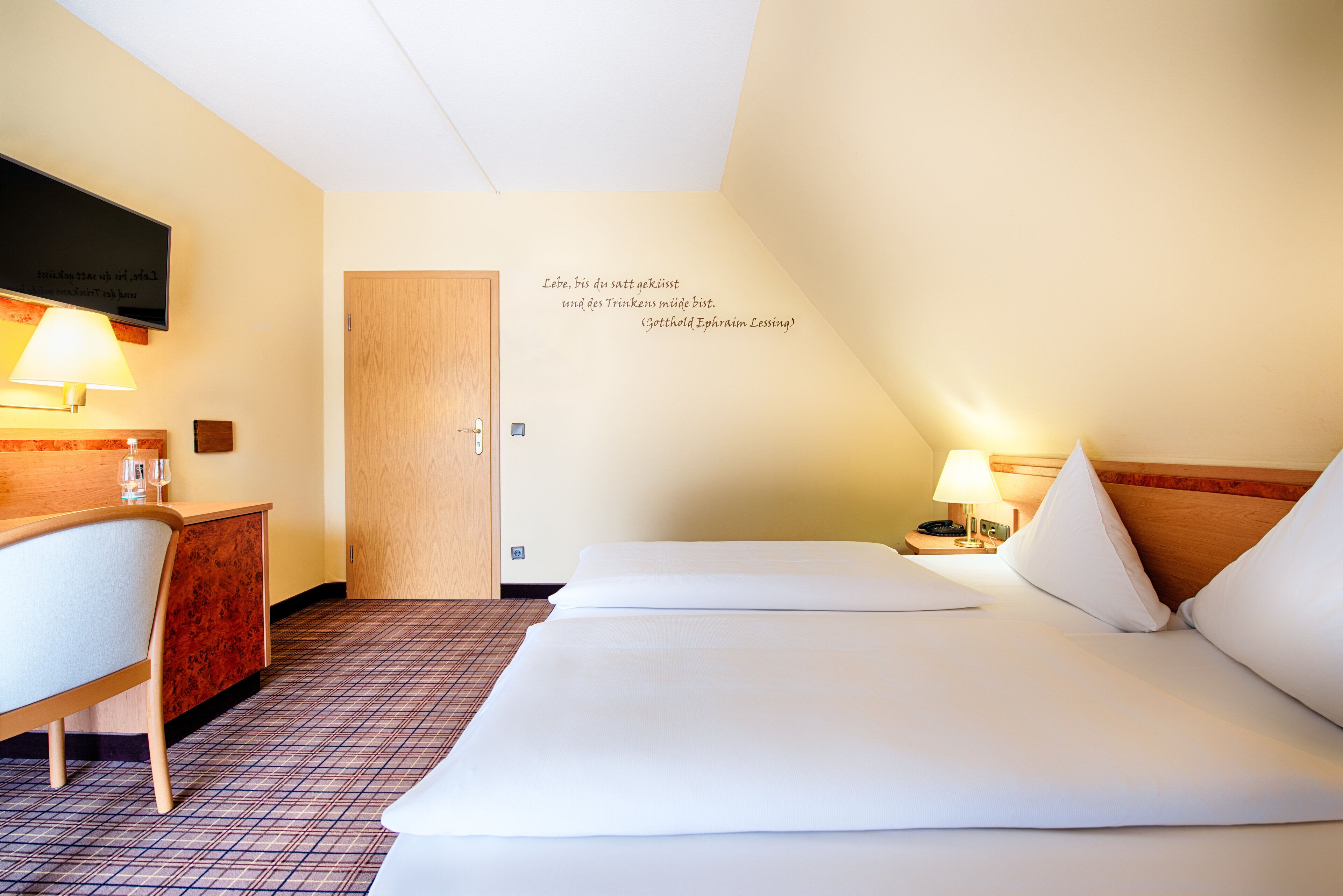A hotel room with a bed, a desk, a chair, a TV, and a wall with a quote.