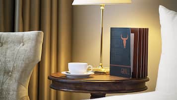 Ein Hotelzimmer mit einem Holztischchen, auf dem eine weiße Tasse und ein Untertasse, eine Lampe und ein Buch mit dem Titel "The Grill" stehen.
