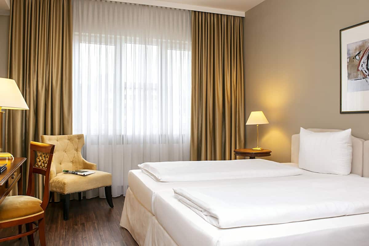 A hotel room with two beds, a desk, a chair, a lamp, and a painting on the wall.