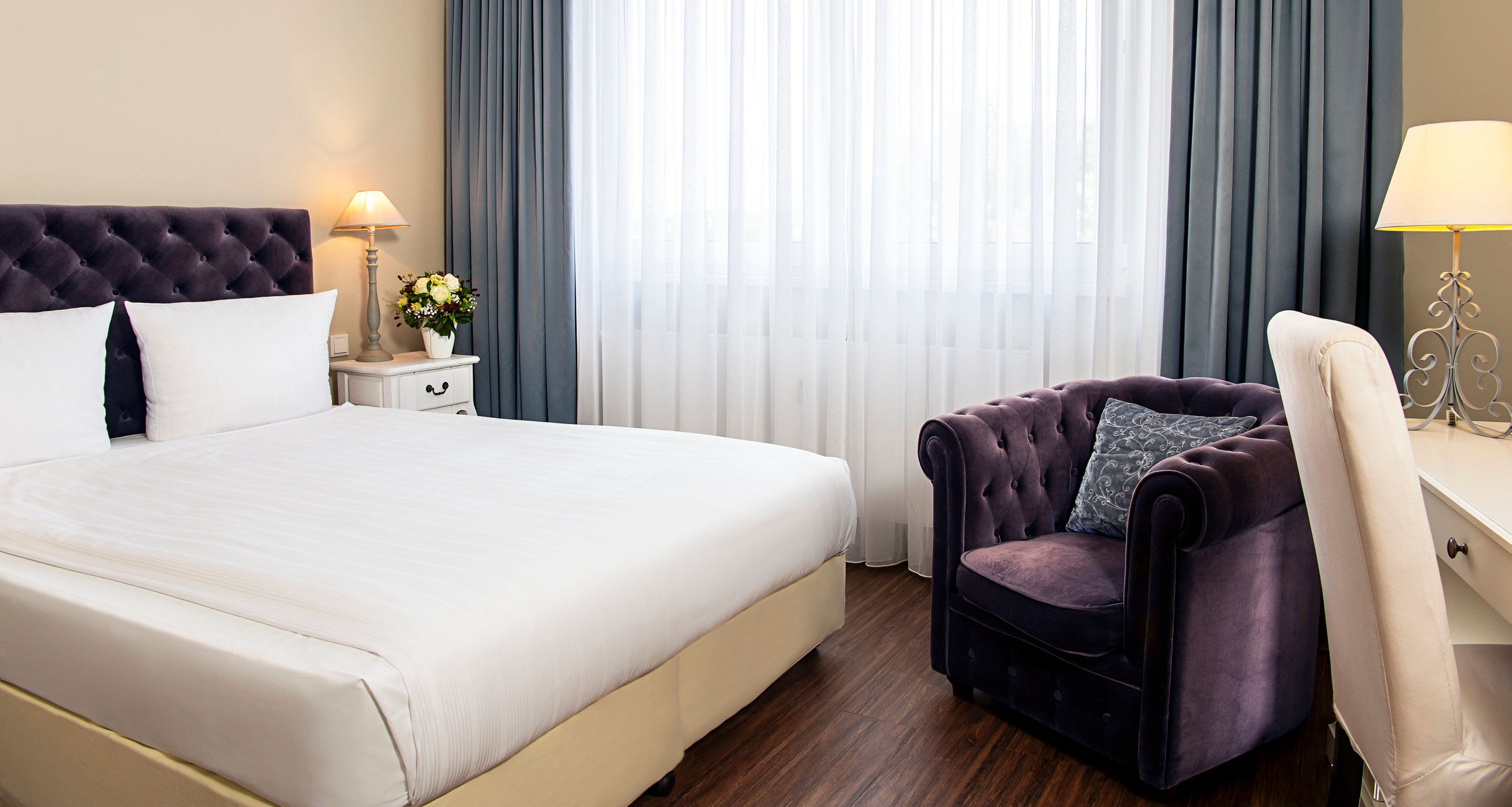 Une chambre d’hôtel avec un grand lit, un fauteuil à revêtement sombre, un bureau blanc et une lampe.