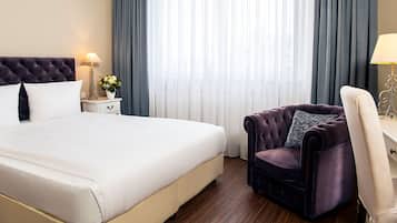 Une chambre d’hôtel avec un grand lit, un fauteuil à revêtement sombre, un bureau blanc et une lampe.