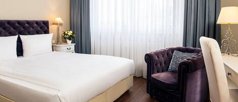 A hotel room with a large bed, a dark upholstered armchair, a white desk, and a lamp.
