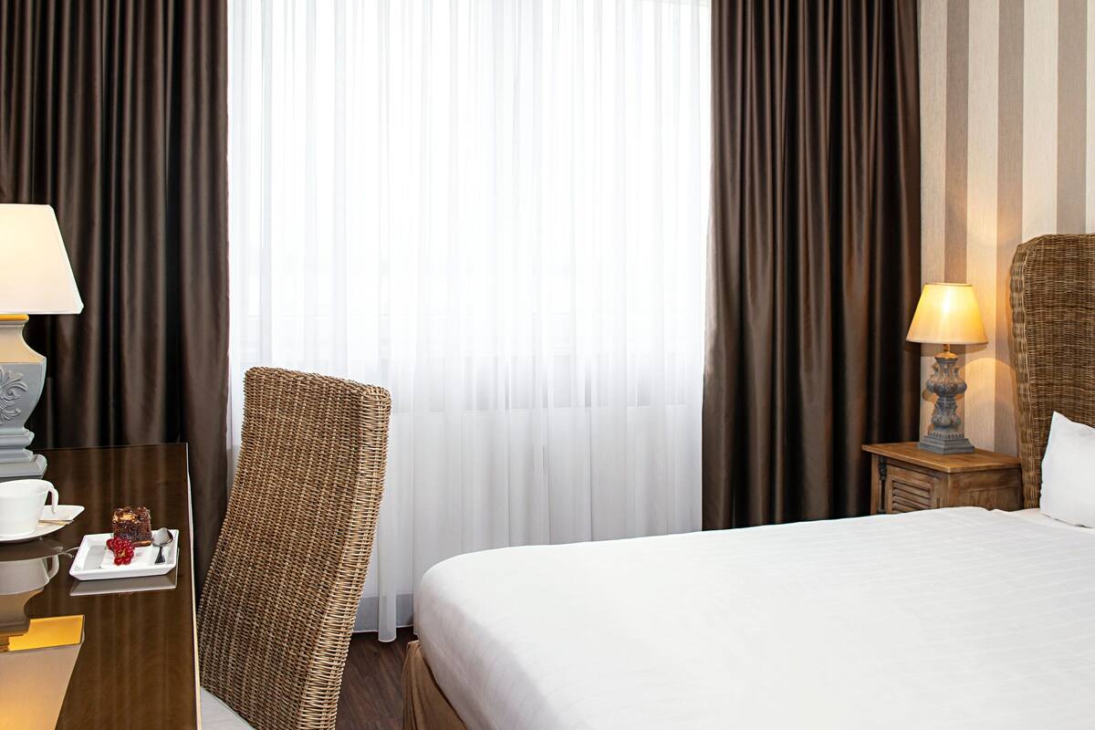 A hotel room with a large bed, a wicker chair, a nightstand with a lamp, and a view of the street through curtains.