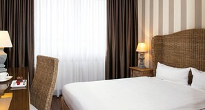 Habitación de hotel con una cama grande, una silla de mimbre, una mesita de noche con lámpara y vista a la calle a través de cortinas.