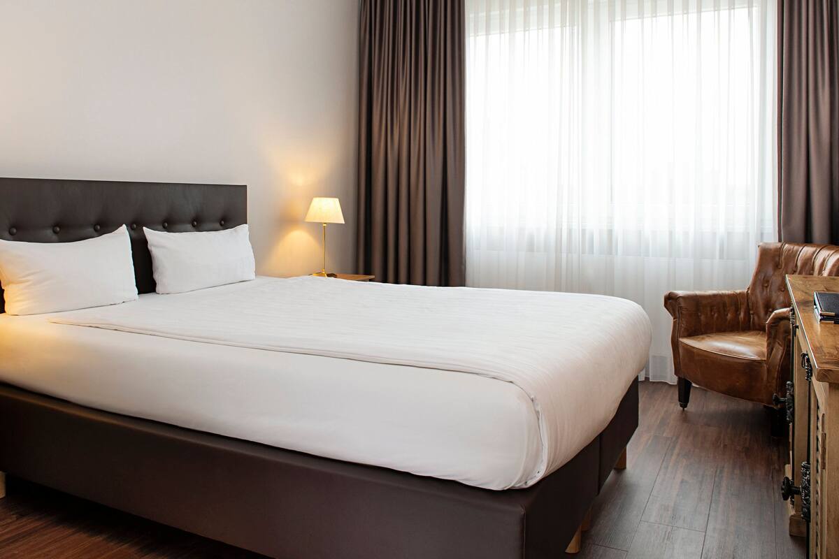 A hotel room with a large bed, two bedside lamps, a wooden nightstand, a leather armchair, and a wooden desk with a laptop.