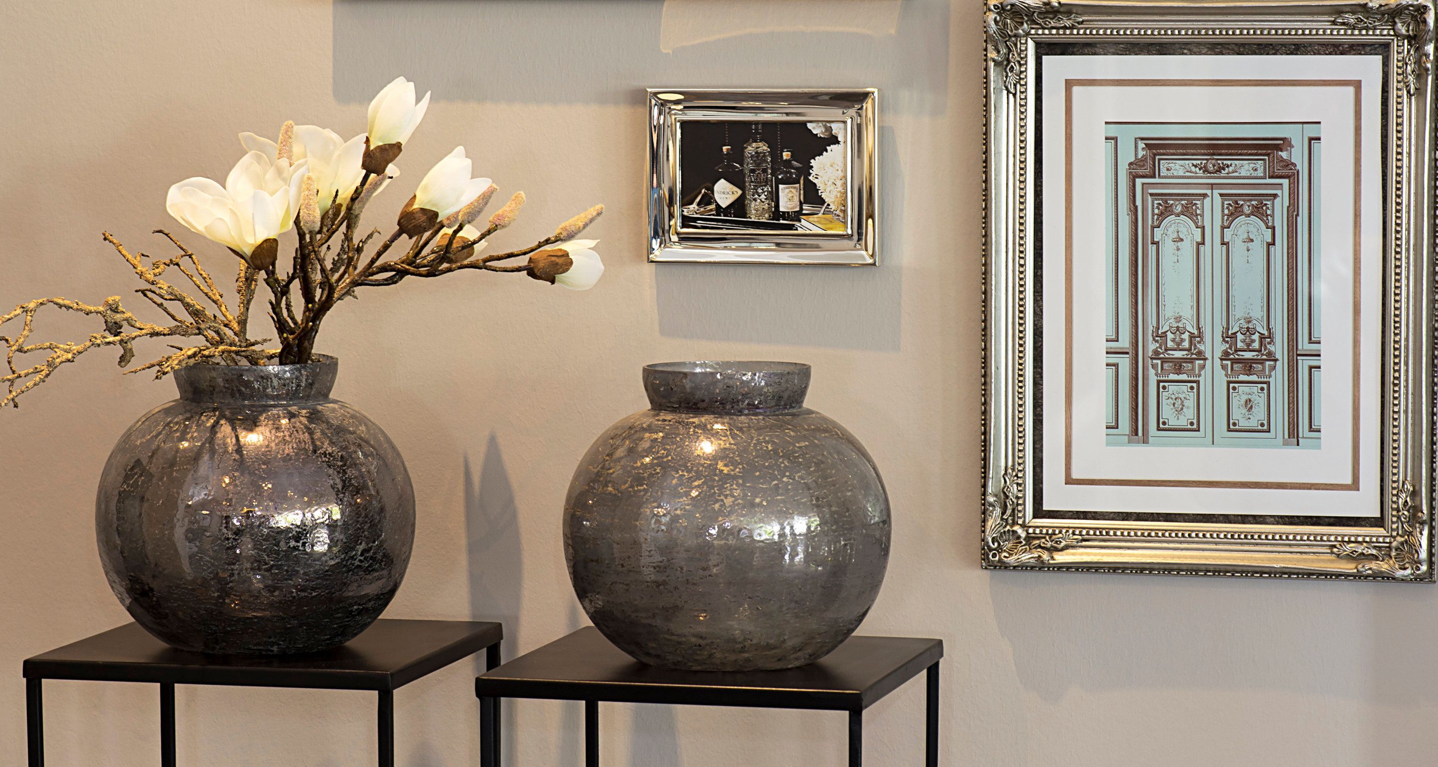 Two vases with flowers on a black pedestal, a framed photograph, and a framed picture of a building on a wall.