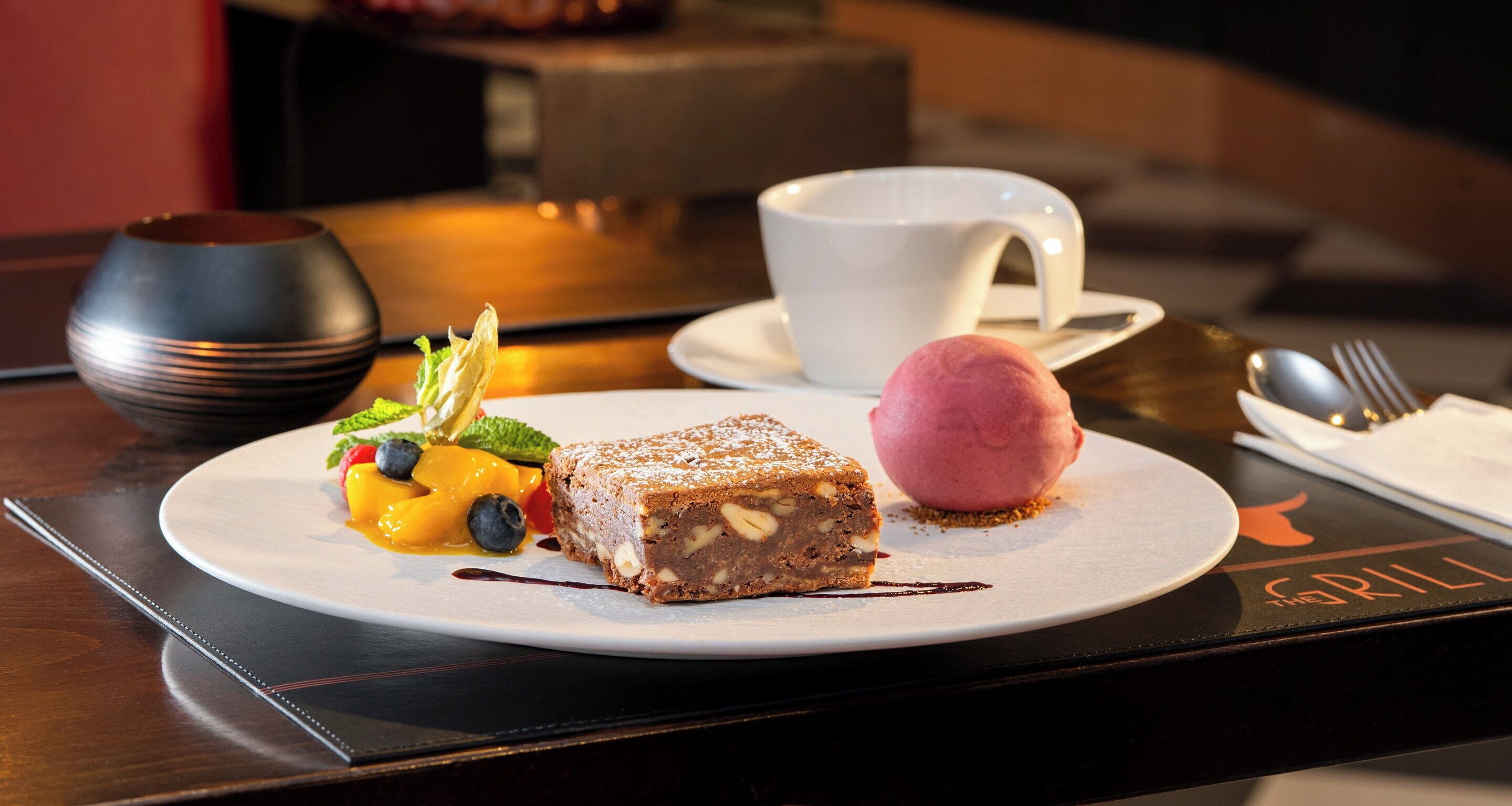 Un plato de postre con una porción de pastel, una bola de helado y fruta fresca sobre un plato blanco.