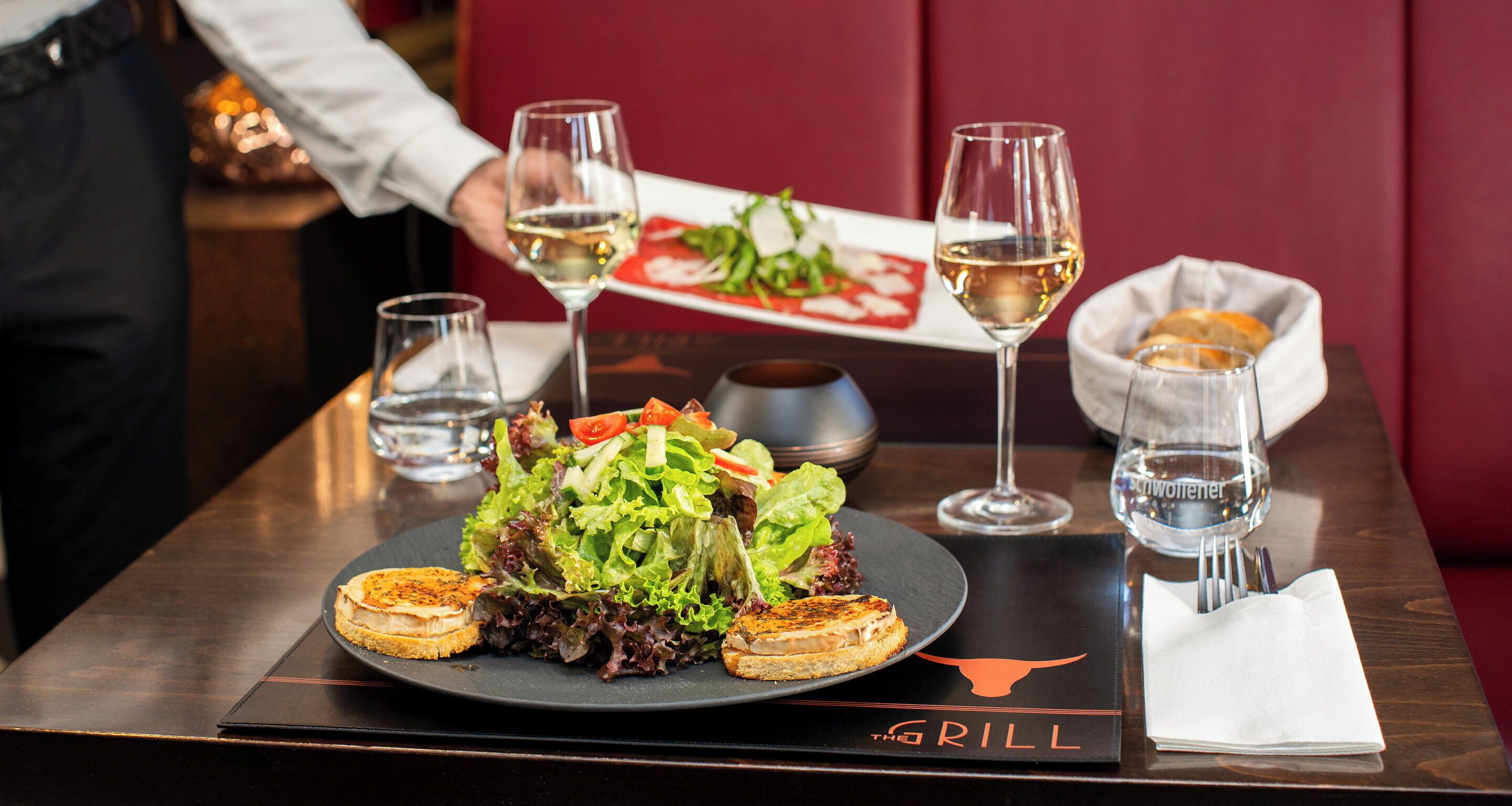 Una mesa de comedor con ensalada, carnes a la parrilla y copas de vino blanco.