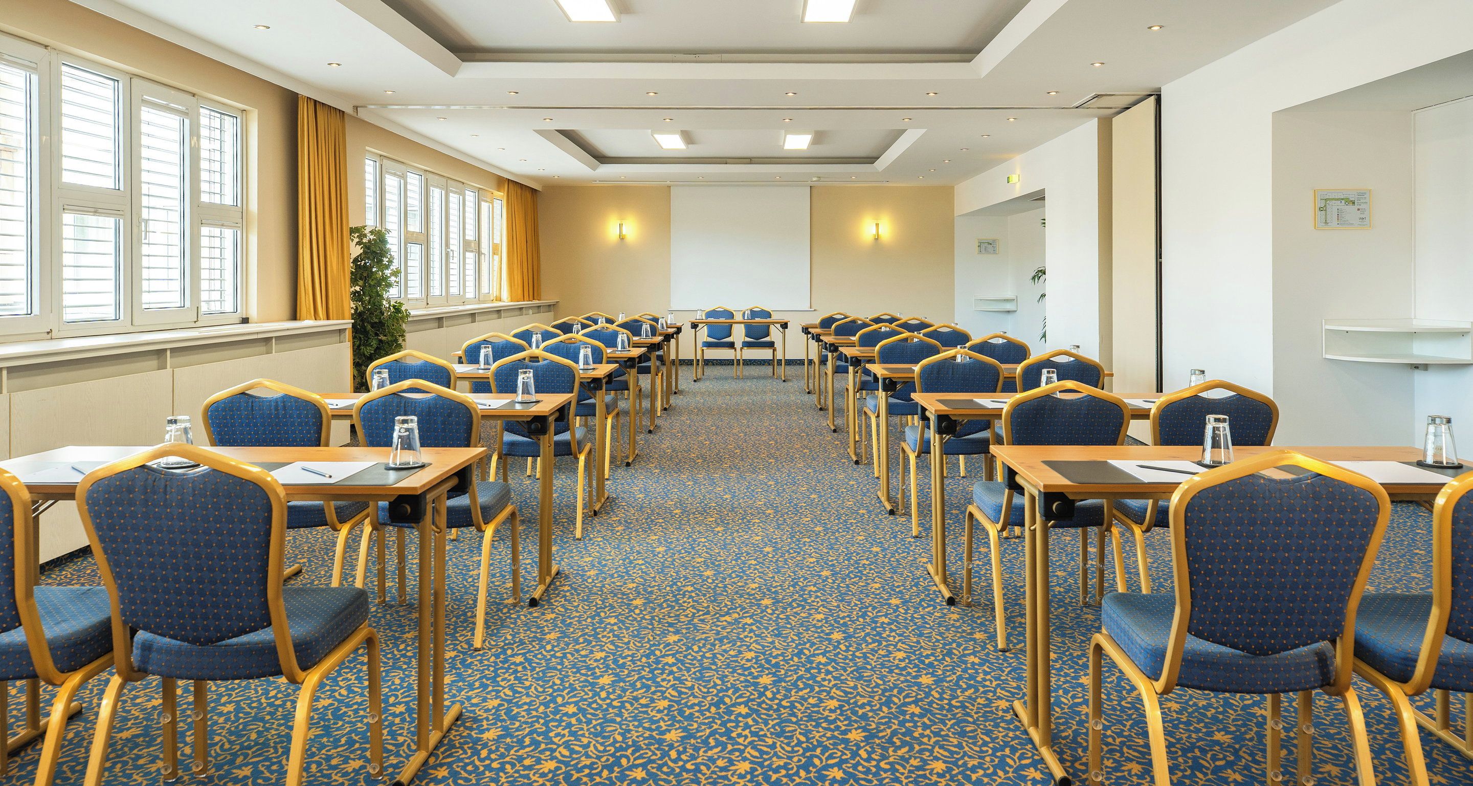 Sala de conferência com mesas e cadeiras dispostas em fileiras, amplas janelas e piso coberto de carpete.