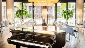 A grand piano in a spacious, well-lit room with large windows, clear chairs, and tables set for dining.