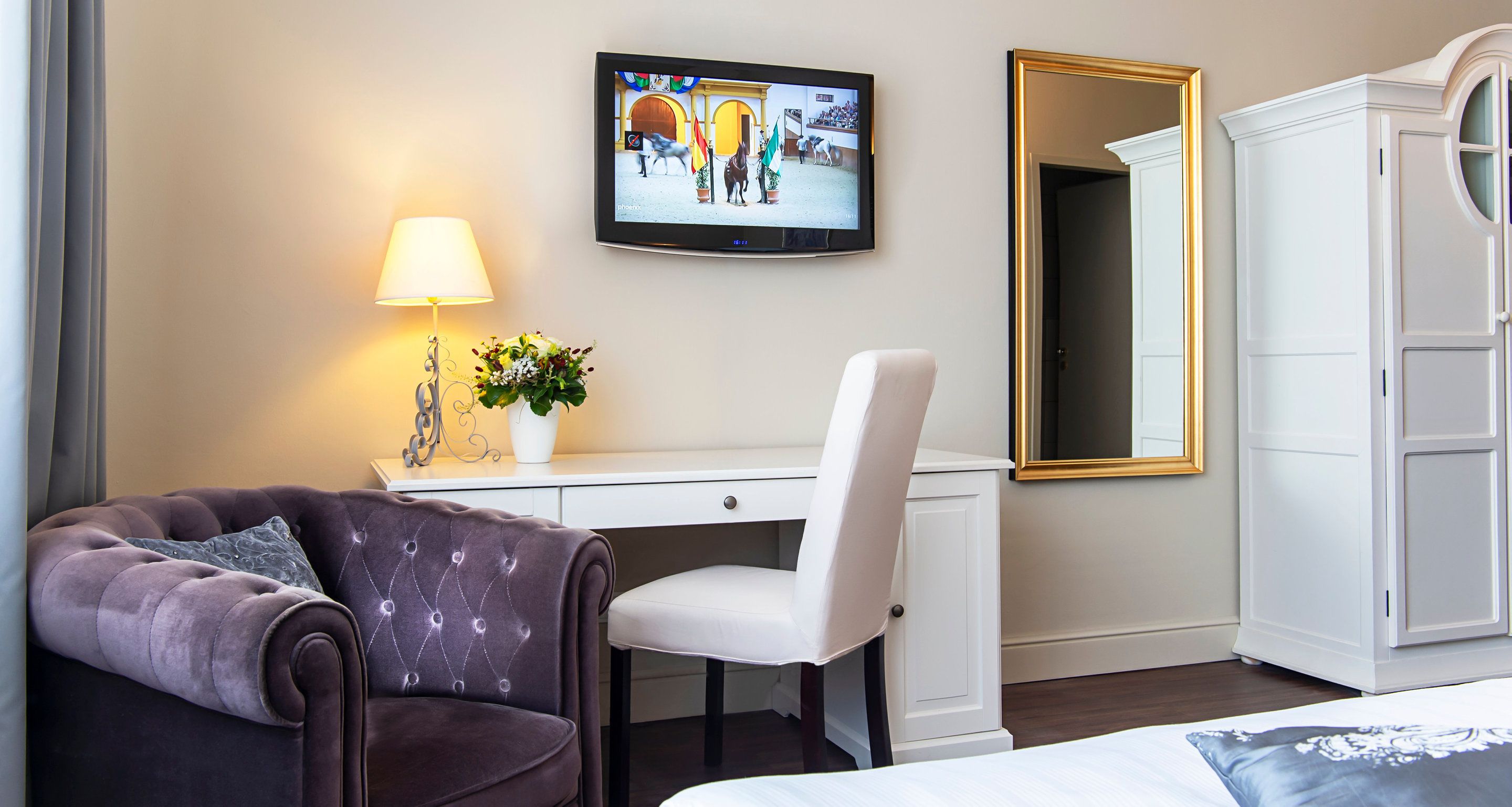 A hotel room with a dark brown sofa, a white desk, a television mounted on the wall, a white wardrobe, and a mirror.
