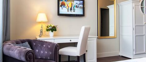 A hotel room with a dark brown sofa, a white desk, a television mounted on the wall, a white wardrobe, and a mirror.