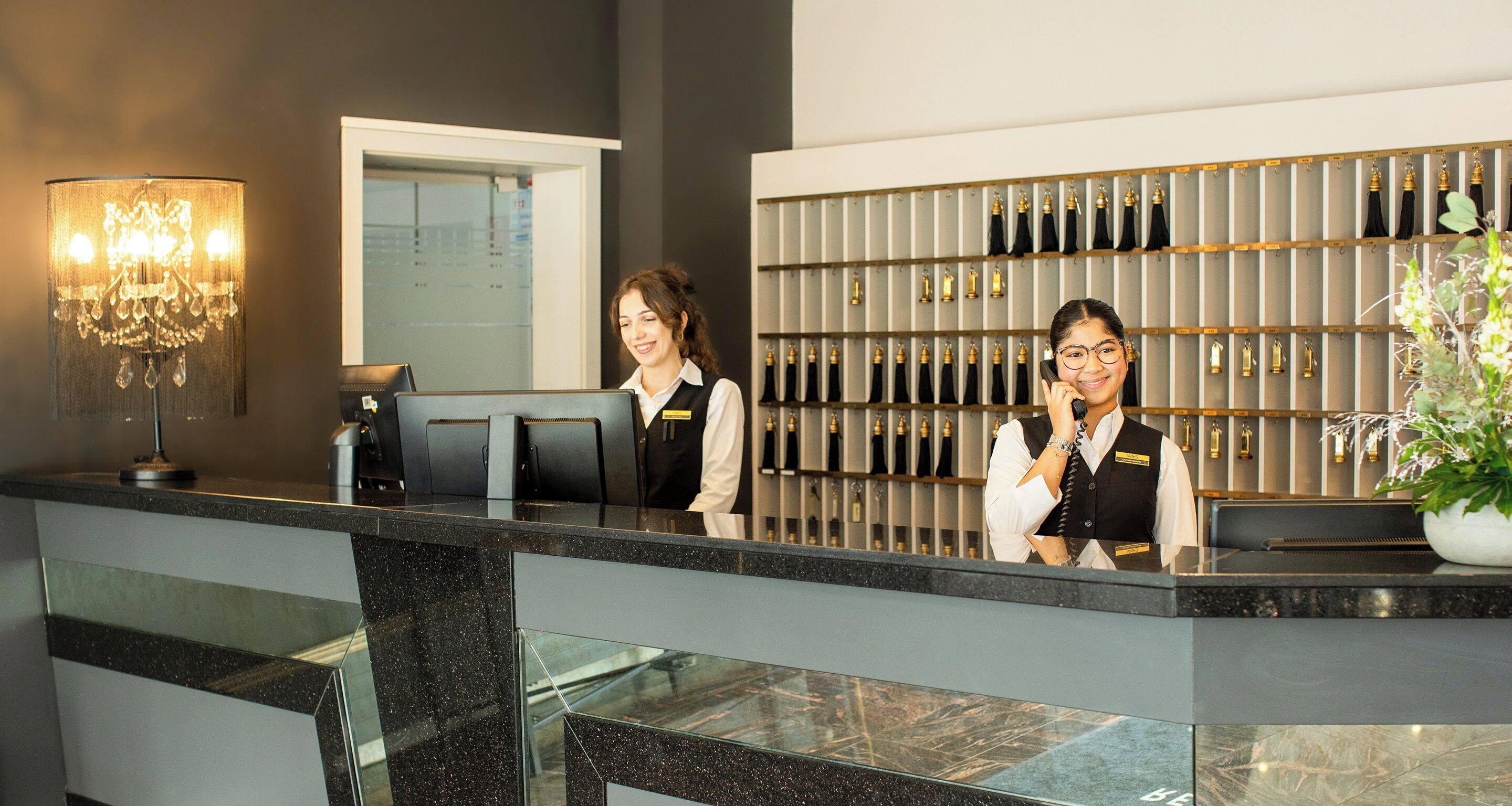 Et hotelreceptionsområde med et moderne design, herunder en receptionsdisk, en lysekrone og en vægmonteret udstilling af flasker.