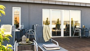 A modern outdoor patio with a wooden deck, metal chairs, a table, and a glass door leading to a balcony.